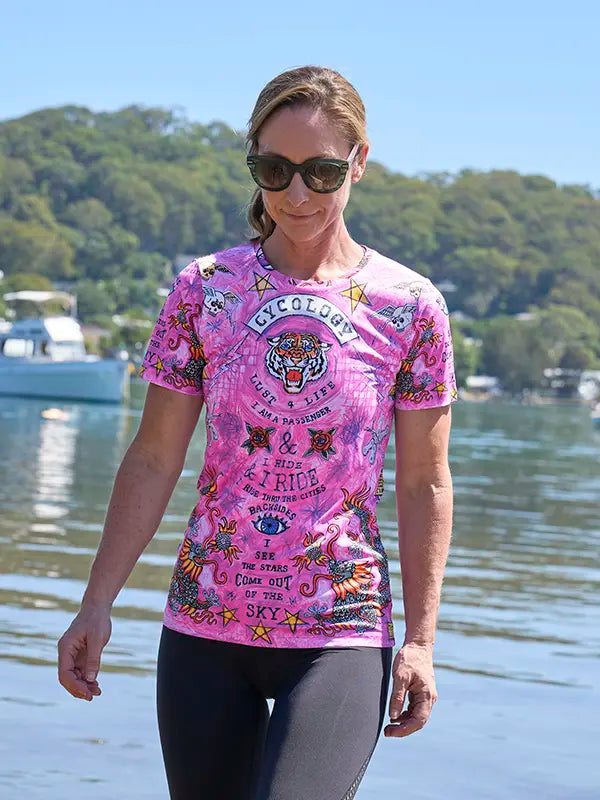 Woman wearing a pink graphic tech t-shirt by a body of water with trees in the background