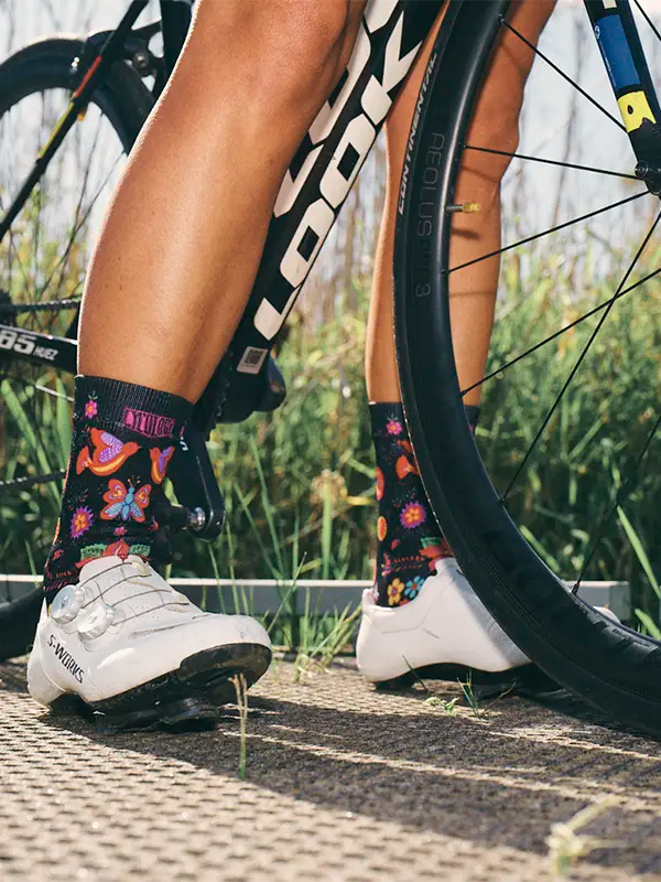 Person wearing cycling shoes and Frida Black socks next to a bicycle on a paved path with grass in the background