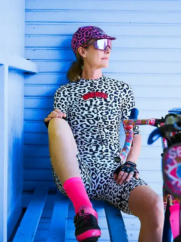 Woman in leopard print cycling kit with pink socks and sunglasses, leaning against blue wall
