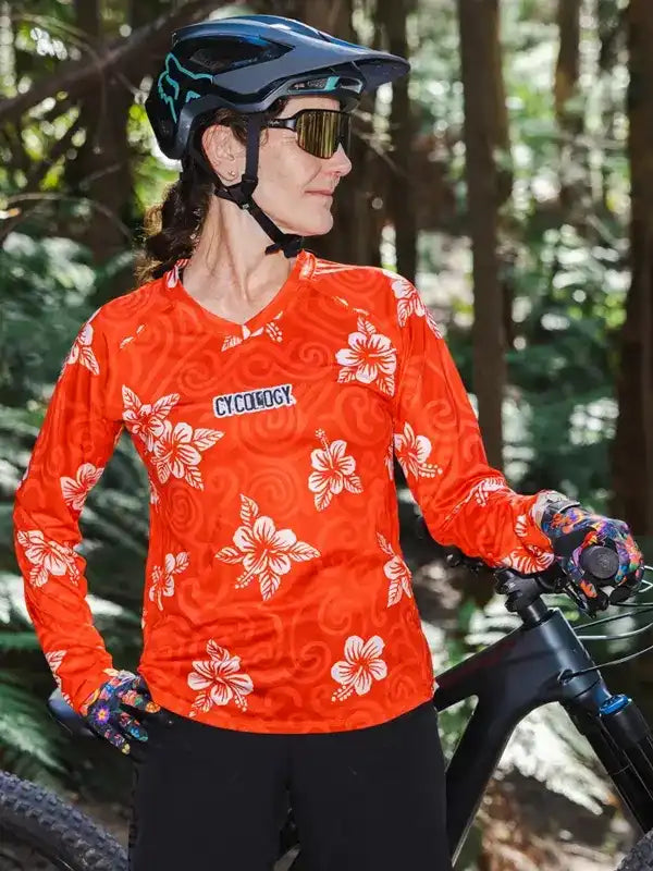 Woman in orange floral cycling jersey with helmet, sunglasses, and gloves, standing with mountain bike in forest