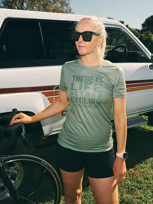 Woman in olive cycling tee with 'There Is Life Beyond Cycling' text, standing by bike and van outdoors