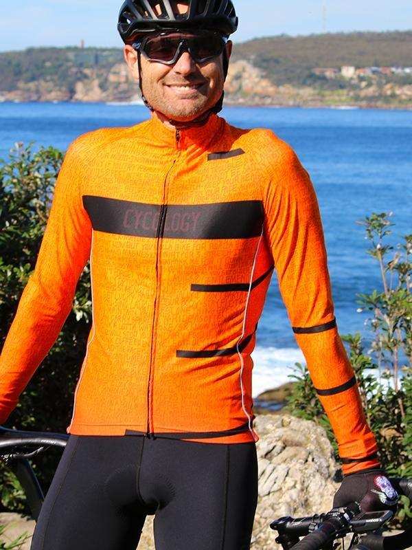 Man in bright orange long-sleeve cycling jersey with black accents, helmet, and sunglasses, by coastal scenery