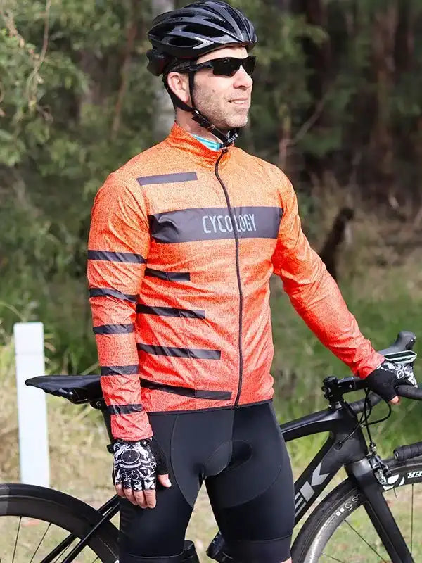 Man in orange cycling jersey with black stripes, helmet, and gloves, standing beside road bike outdoors