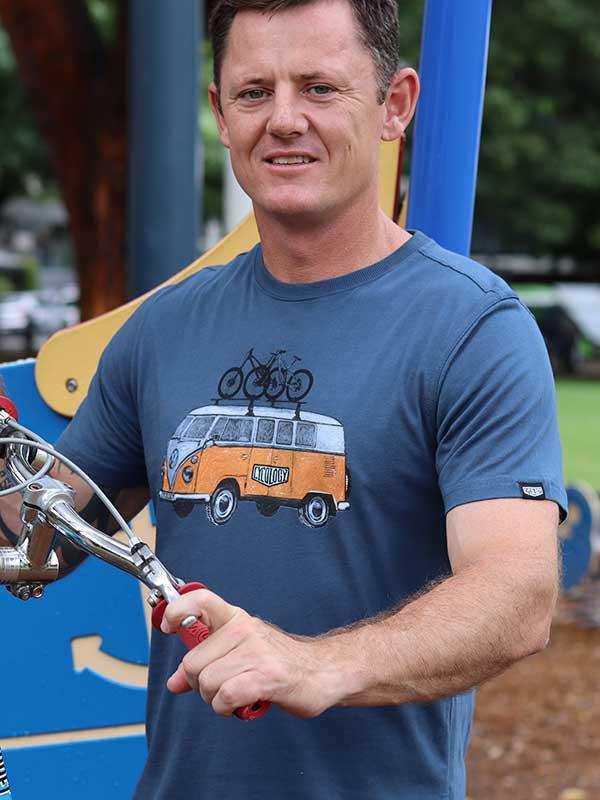 Man wearing blue cycling t-shirt with vintage van graphic, holding bike handlebars outdoors