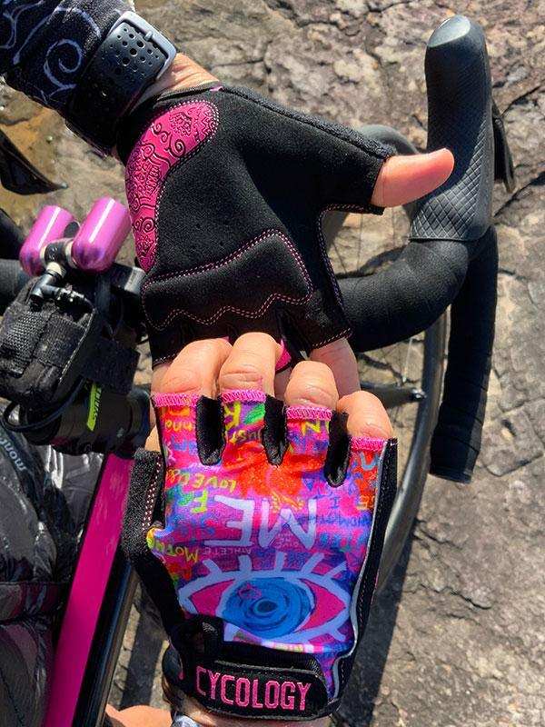 Colorful graffiti-style cycling gloves with pink accents, worn on bike handlebars, featuring fingerless design and brand tag.