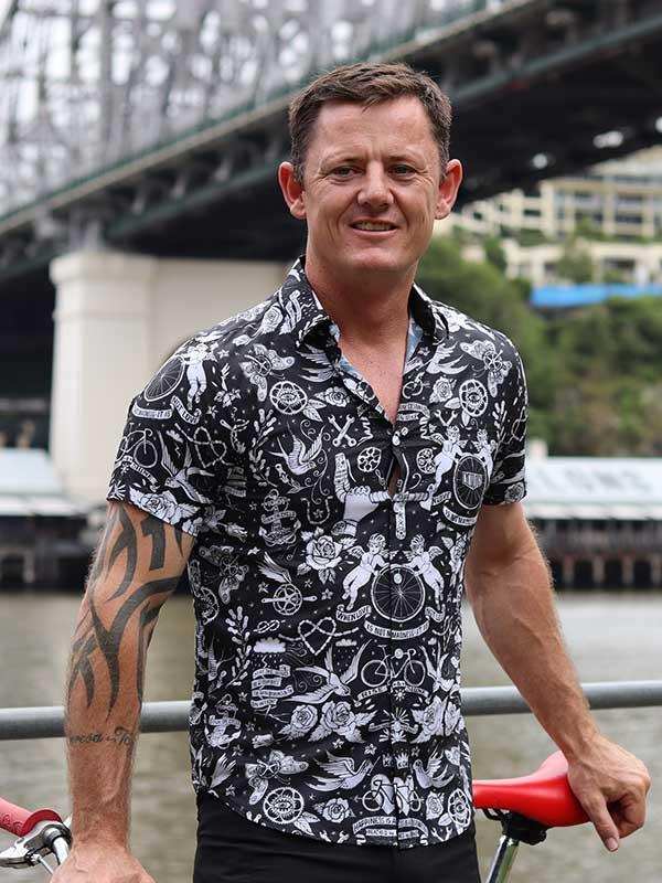 Man wearing black and white cycling-themed shirt, standing by river with bridge, holding red bike