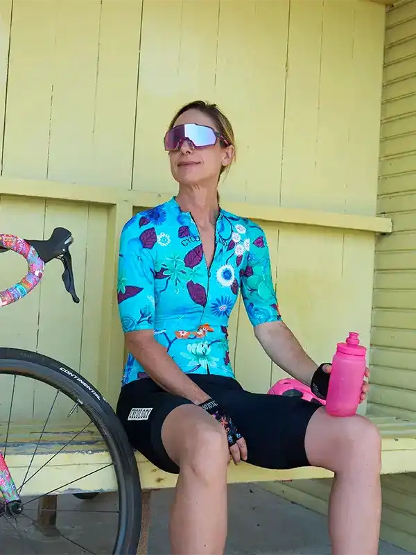 Woman in floral cycling jersey and sunglasses holding pink water bottle beside bike