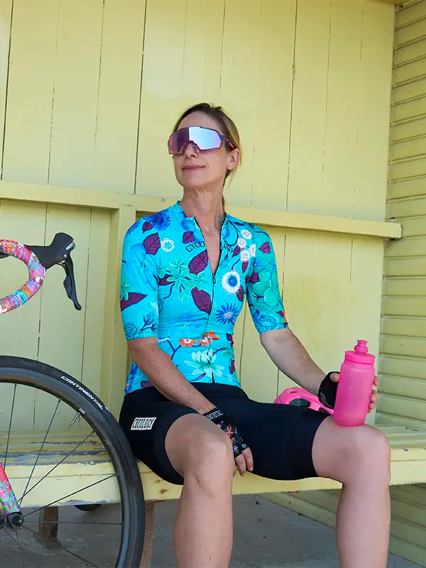Person wearing  Aloha Aqua Women's Reborn Cycling Jersey cycling gear sitting on a bench next to a bicycle