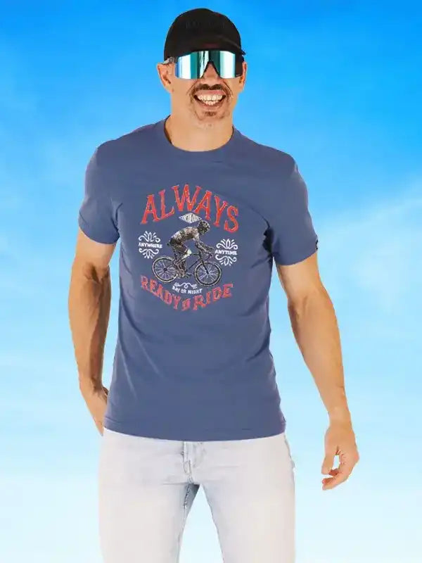 Man wearing blue cycling t-shirt with 'Always Ready to Ride' graphic, sunglasses, and cap against blue sky