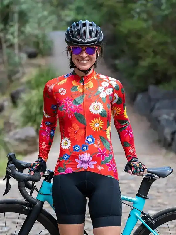 Woman in vibrant floral cycling jersey and black bib shorts, holding turquoise road bike on forest trail