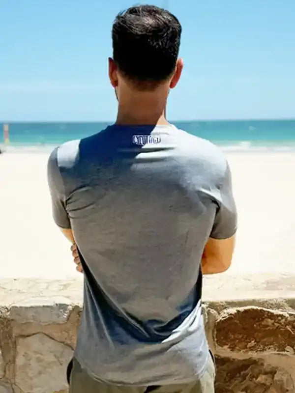Man in gray cycling jersey with blue back panel, standing by beach, ideal for men's cycling apparel