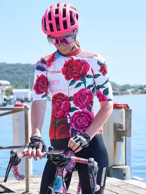Woman in floral cycling jersey and pink helmet on dock with bike, seaside backdrop