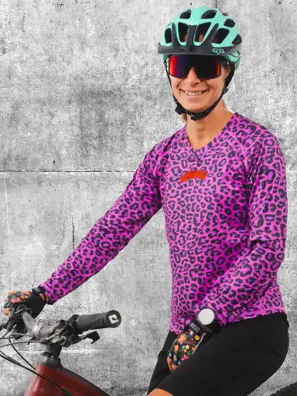 Woman in vibrant purple leopard print cycling jersey, helmet, and sunglasses, riding bike against concrete wall