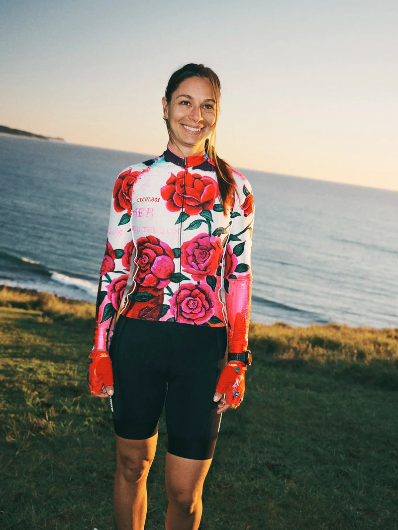 Woman in floral long sleeve cycling jersey and black shorts, standing by ocean at sunset