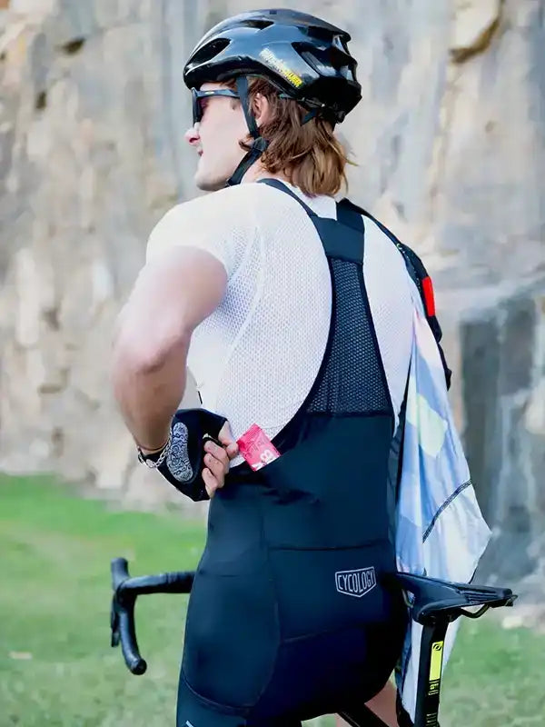 Man in black cycling bib shorts and white jersey, wearing helmet, standing beside road bike outdoors