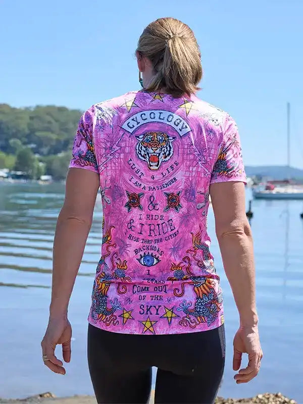 Pink women's cycling jersey with tiger graphic, stars, and 'I ride & I ride' text, worn by cyclist near water.