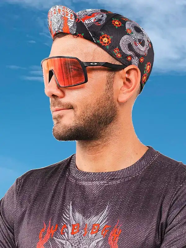 Man wearing cycling bandana with skull and floral design, sunglasses, and performance tee under blue sky