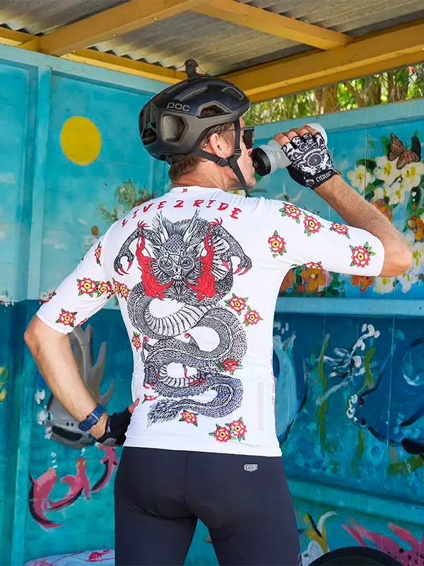 Person wearing a white cycling jersey with a dragon design, black helmet, and gloves, standing in front of a colorful wall.