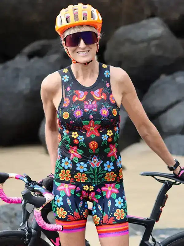 Woman in colorful floral cycling jersey and shorts with orange helmet, standing beside road bike on rocky beach