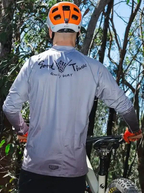 Cyclist wearing gray long-sleeve jersey with 'Good Times Quality Dirt' graphic, orange helmet, and gloves on trail