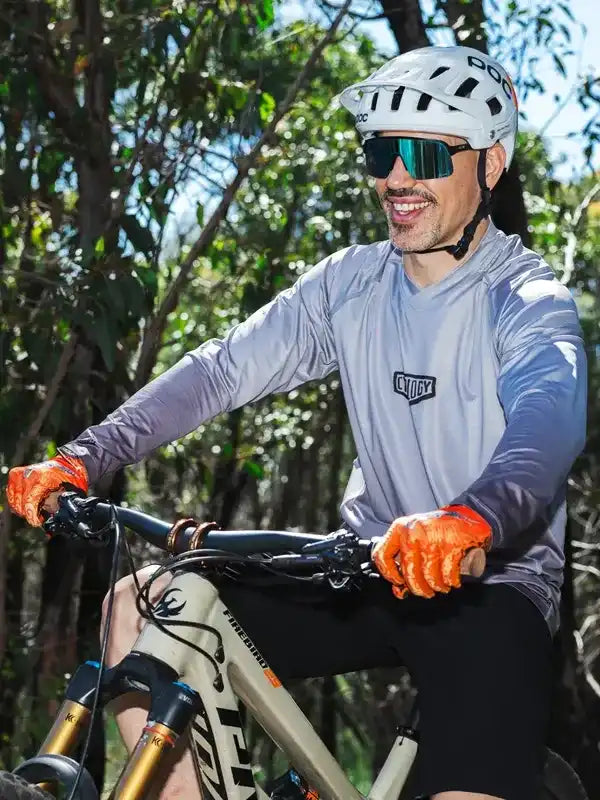 Man cycling in forest wearing gray long-sleeve jersey, orange gloves, and white helmet