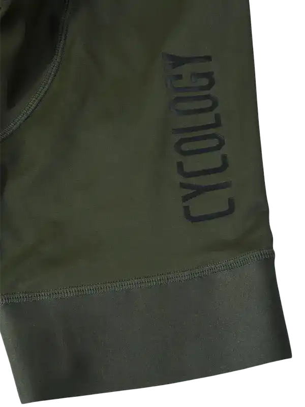 Close-up of an olive green bib short with a dark gray vertical 'CYCOLOGY' logo and a ribbed cuff, 
