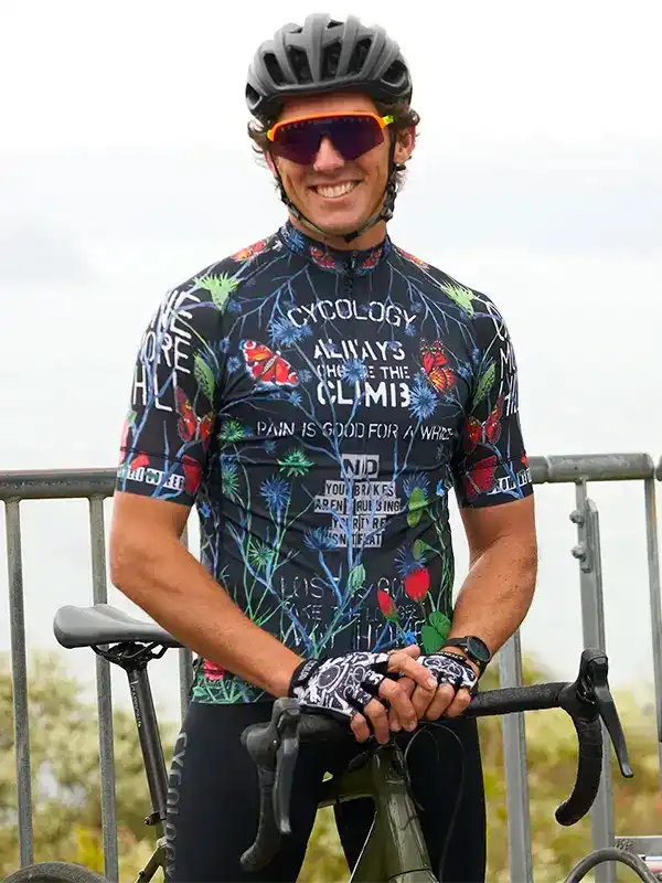 Man in floral cycling jersey with 'Always Choose the Climb' slogan, helmet, and sunglasses, holding bike outdoors