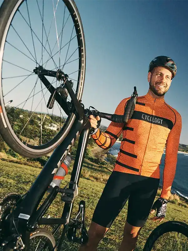 Man in orange cycling jersey and black shorts holding bike outdoors, wearing Cycology gear
