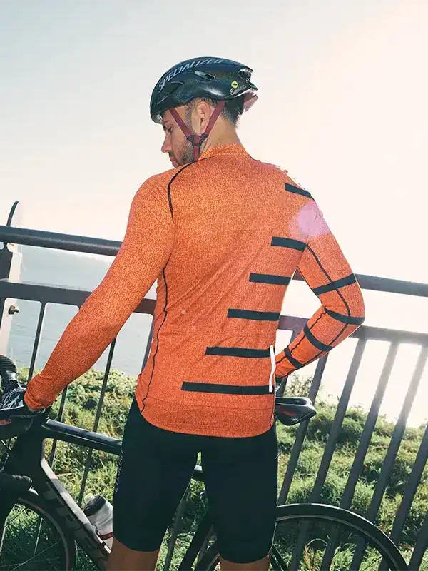 Man in orange long-sleeve cycling jersey with black stripes, wearing helmet, standing by bike outdoors