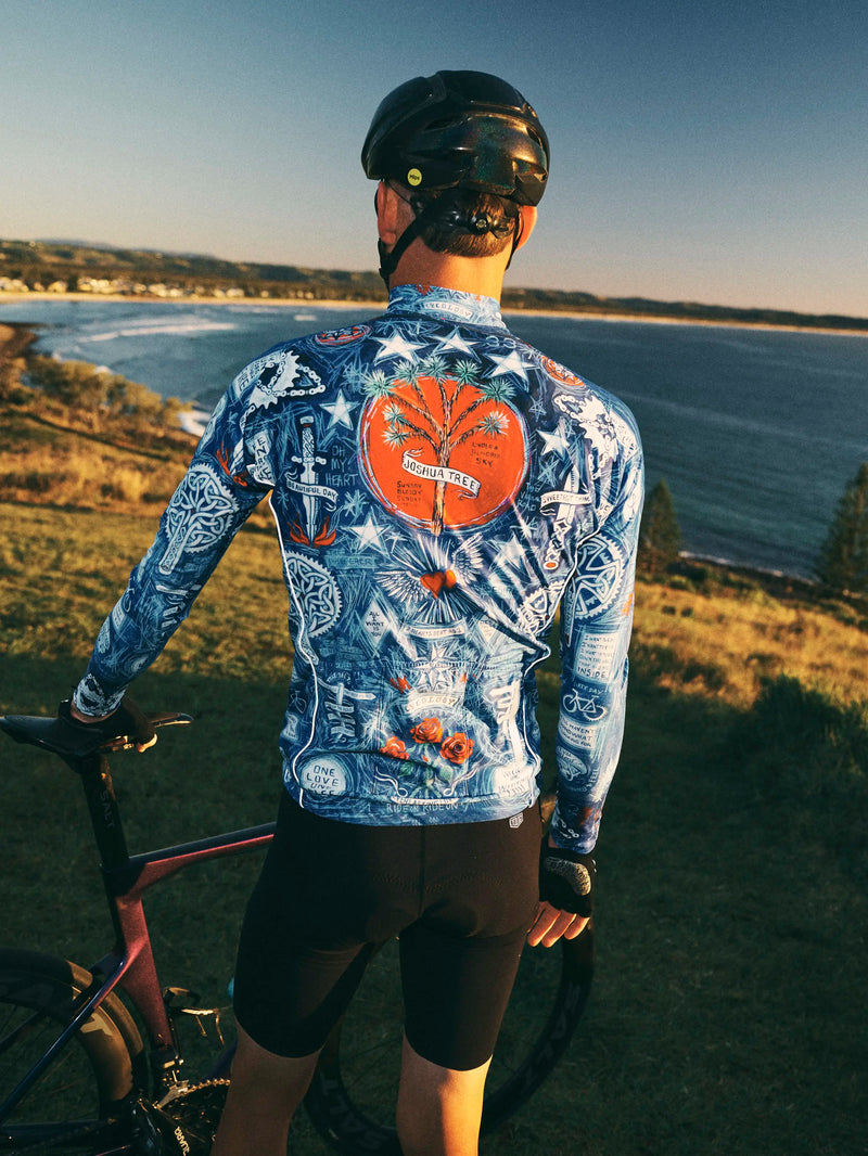 Man in a colorful blue cycling Long Sleeve Jersey standing next to a bicycle by the sea.