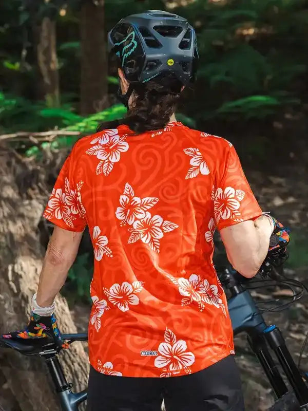Woman in vibrant orange floral cycling jersey riding mountain bike through forest trail