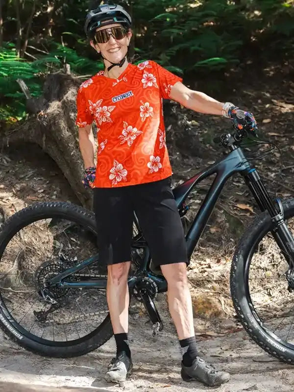 Woman in red floral cycling jersey and black shorts standing with mountain bike on forest trail