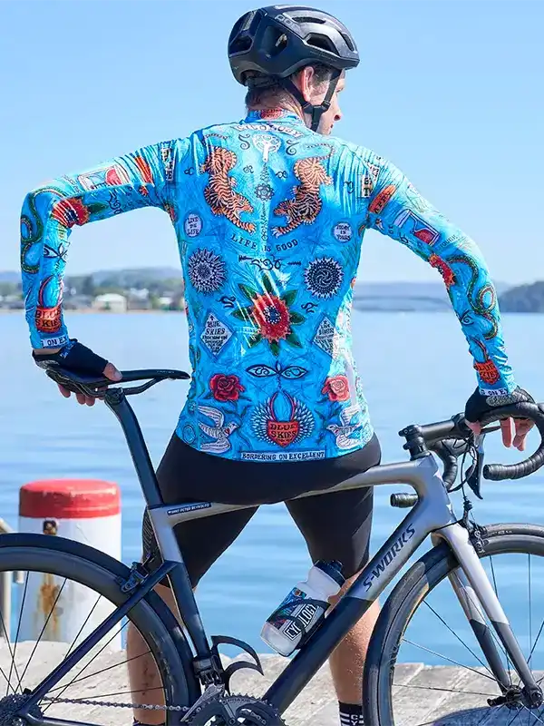 Cyclist in vibrant blue long-sleeve jersey with tiger and rose print, standing by water with bike