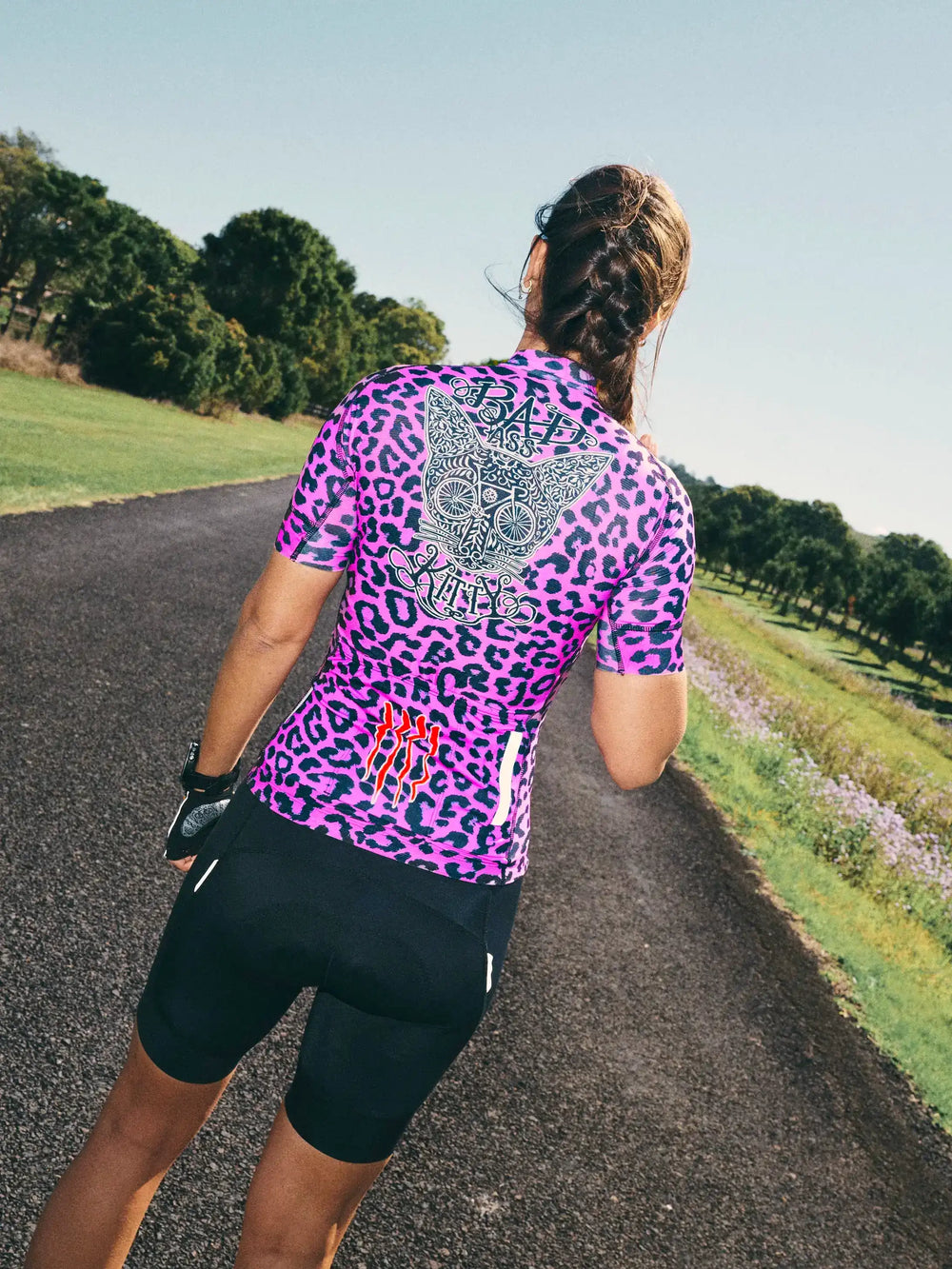 Woman cycling in pink leopard print jersey with 'Bad Ass Kitty' graphic, black shorts, on rural road