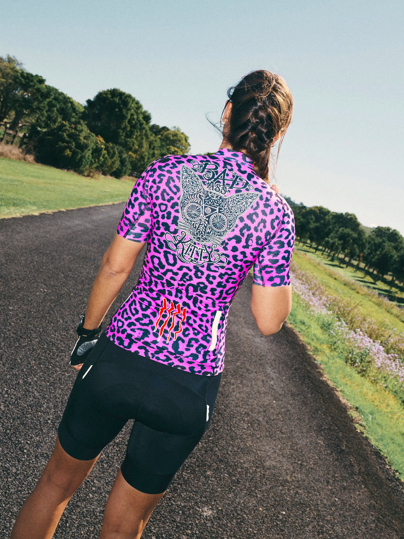 Person wearing a pink leopard print cycling jersey on a road with greenery.