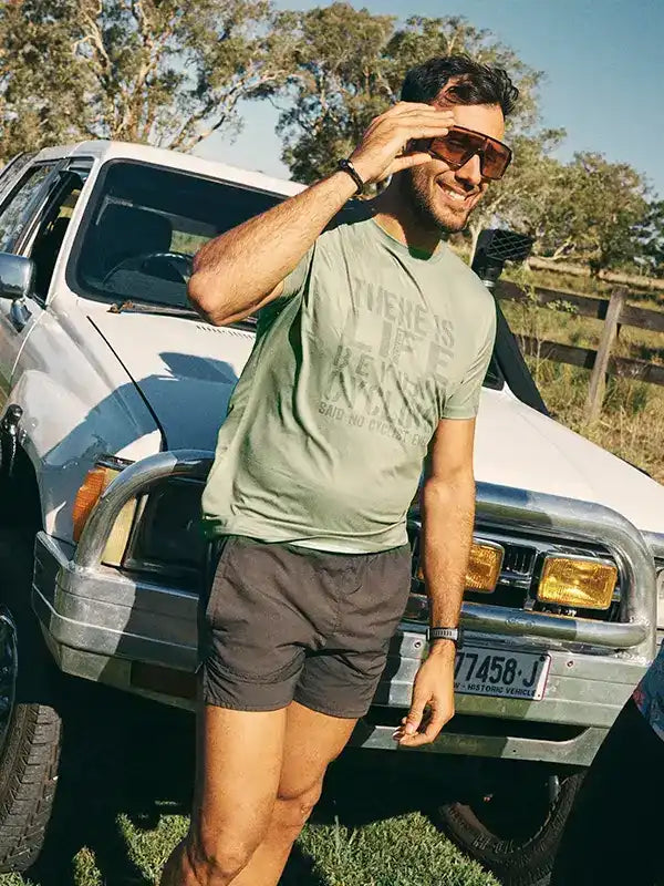 Man in olive cycling tee and shorts leaning on truck, outdoor adventure style