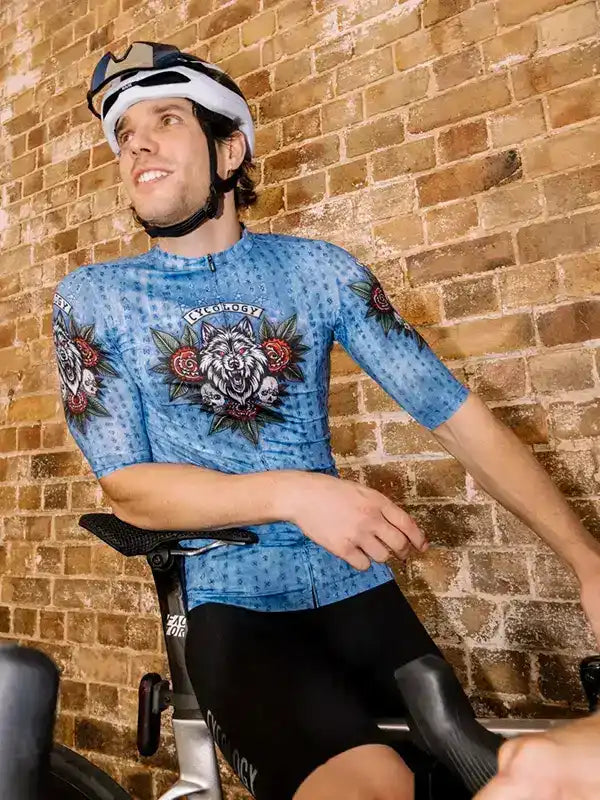 Man wearing blue cycling jersey with wolf and rose design, black shorts, helmet, on stationary bike against brick wall