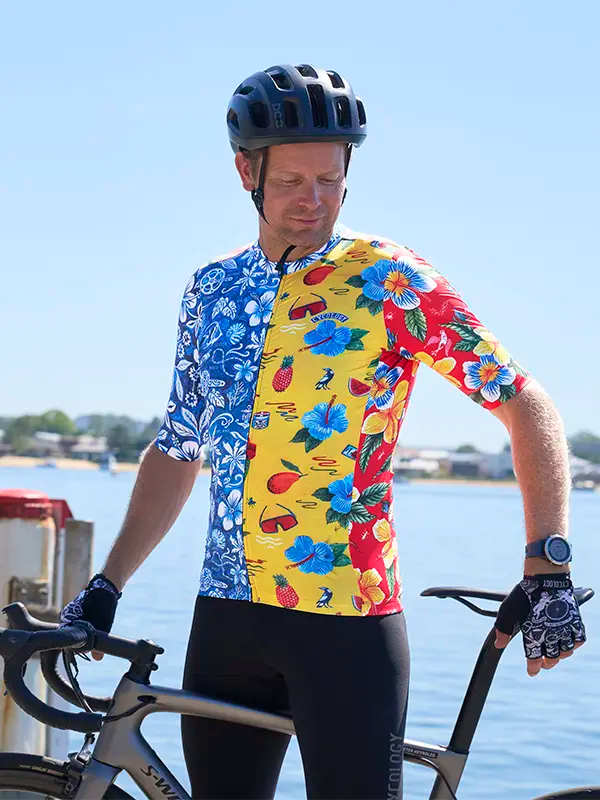 Person wearing a colorful Mahalo men's cycling jersey with a floral pattern, standing next to a bicycle by a waterfront.