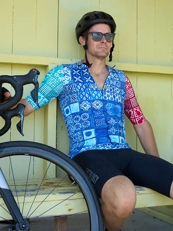 Person wearing a Majorca Men's Blue Reborn  Short Sleeve Cycling Jersey Cycology and helmet, leaning against a yellow wall with a bicycle.