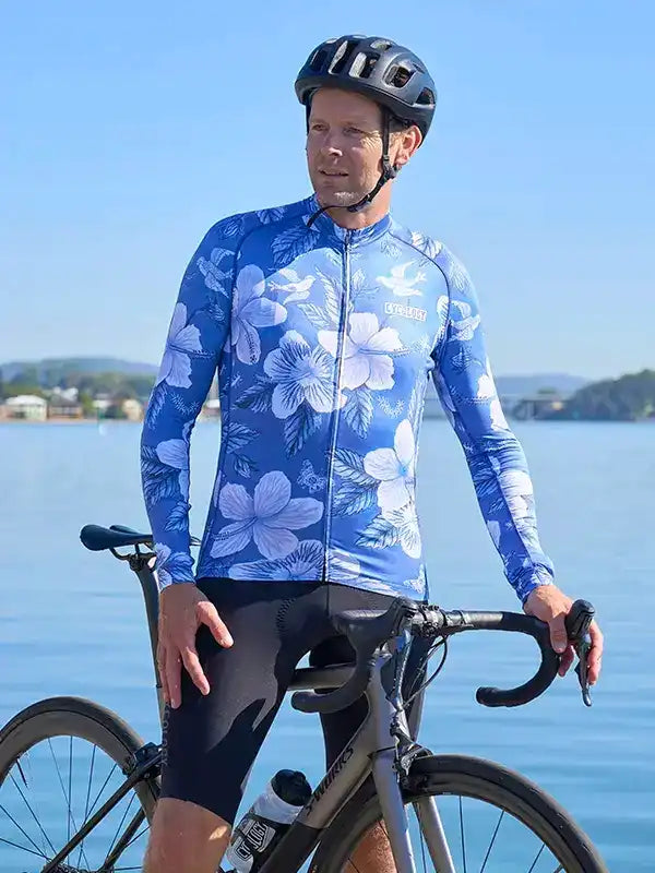 Man in blue floral cycling jersey and black shorts, standing with road bike by water, wearing helmet
