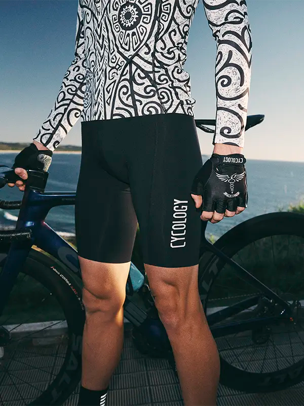 Person wearing black cycling shorts with 'Cycology' branding, standing next to a bicycle by a body of water.