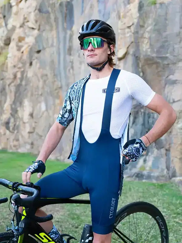 Man in blue cycling bib shorts and white jersey with patterned sleeves, wearing helmet and sunglasses, posing with road bike outdoors