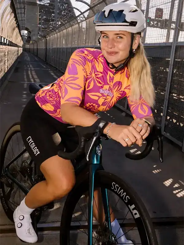 Woman in vibrant floral cycling jersey and black shorts riding bike on bridge, wearing helmet and white shoes