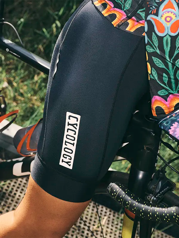 Close-up of a black cycling quick pee bib shorts with 'CYCLOLOGY' branding on a bike seat background.