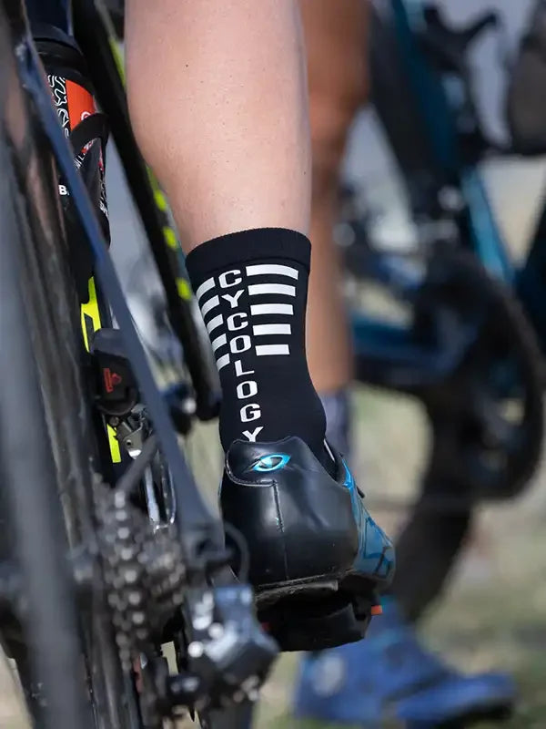 Black cycling sock with white 'CYCOLOGY' text and stripes, worn with clip-in bike shoe on road bike