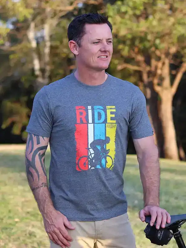 Man wearing gray cycling t-shirt with colorful 'RIDE' graphic, standing outdoors near bike