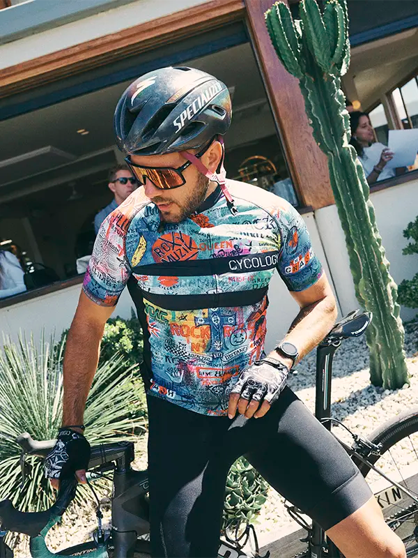 Cyclist in colorful jersey and helmet standing next to a bicycle with a cactus plant in the background.