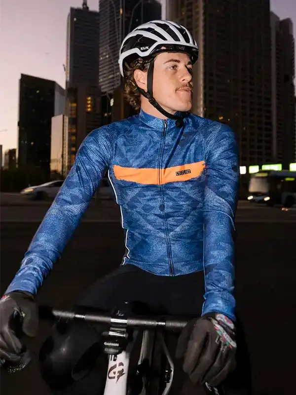 Man in blue patterned cycling jersey with orange chest panel, helmet, gloves, urban skyline backdrop