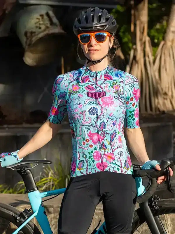 Woman in colorful floral cycling jersey and black shorts, wearing helmet and sunglasses, standing with bike