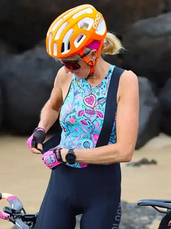 Woman cyclist in colorful patterned jersey, orange helmet, and pink gloves adjusting gear on beach
